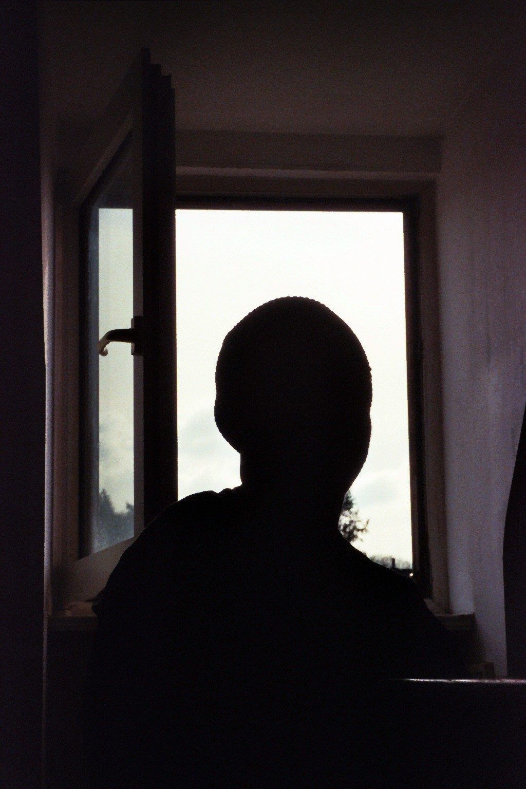 Film photograph: a person silhouetted against an open window, bright overcast sky outside