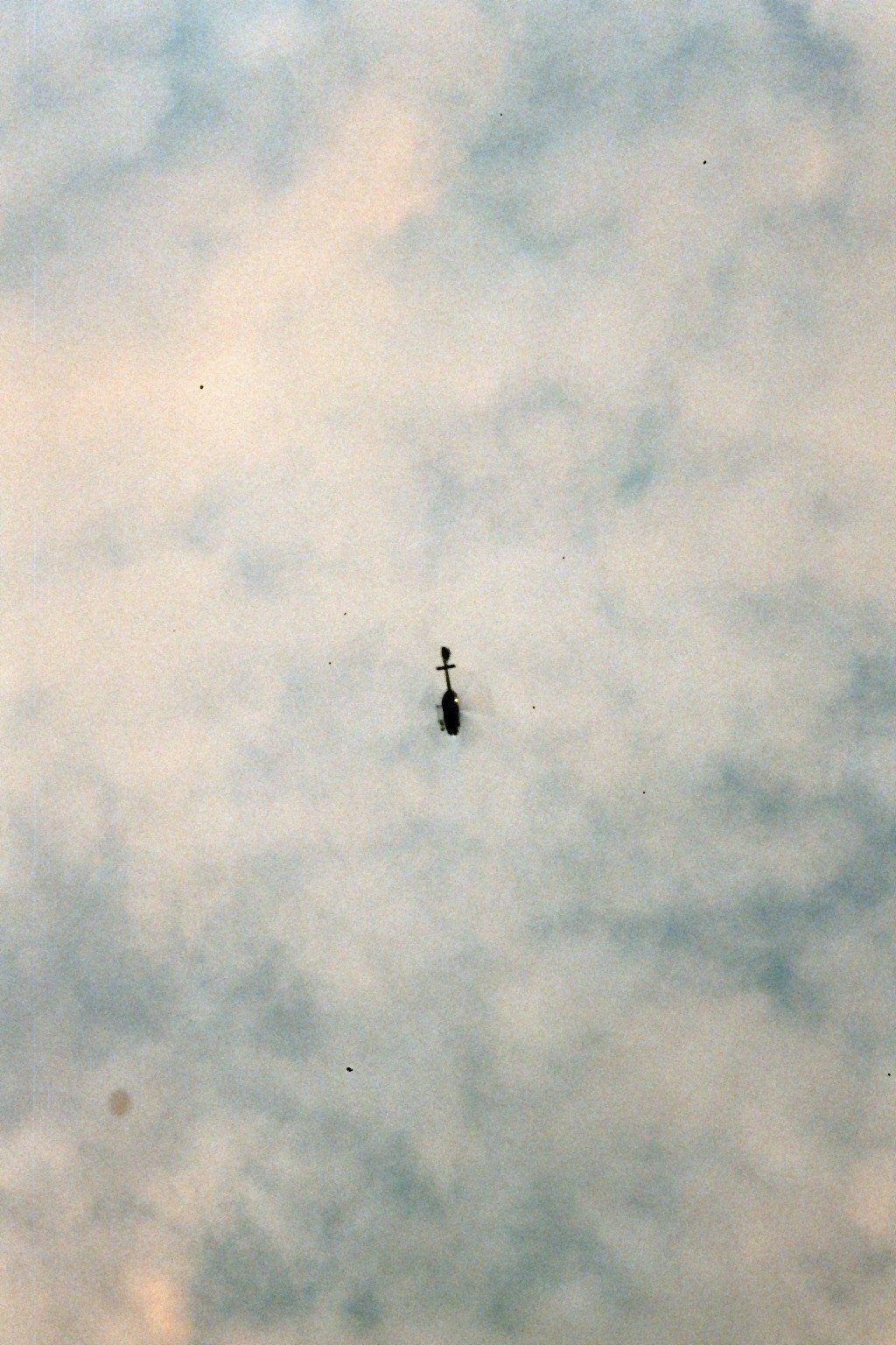 Film photograph: a small helicopter far up in a soft, clouded sky