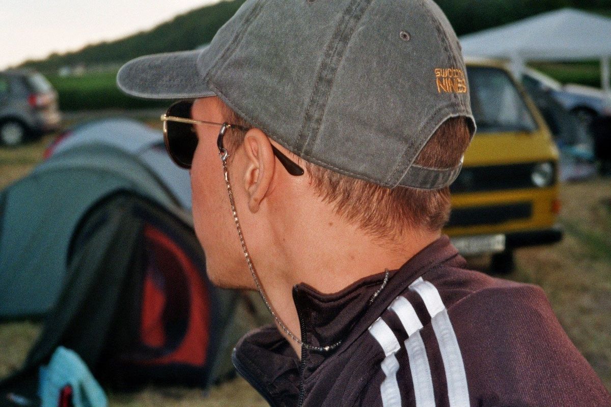 Film photograph: a person in a cap and sunglasses at a campsite, a yellow van and tents in the background