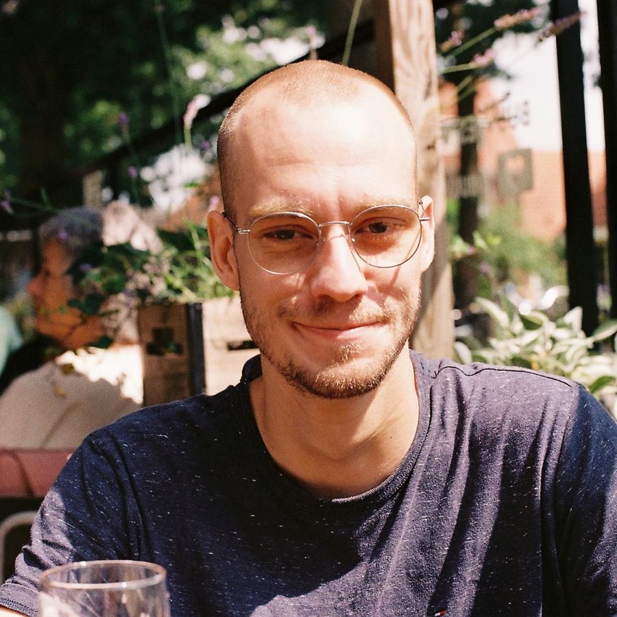 Portrait photograph: the artist sitting outdoors at a café table, wearing glasses, smiling into the camera